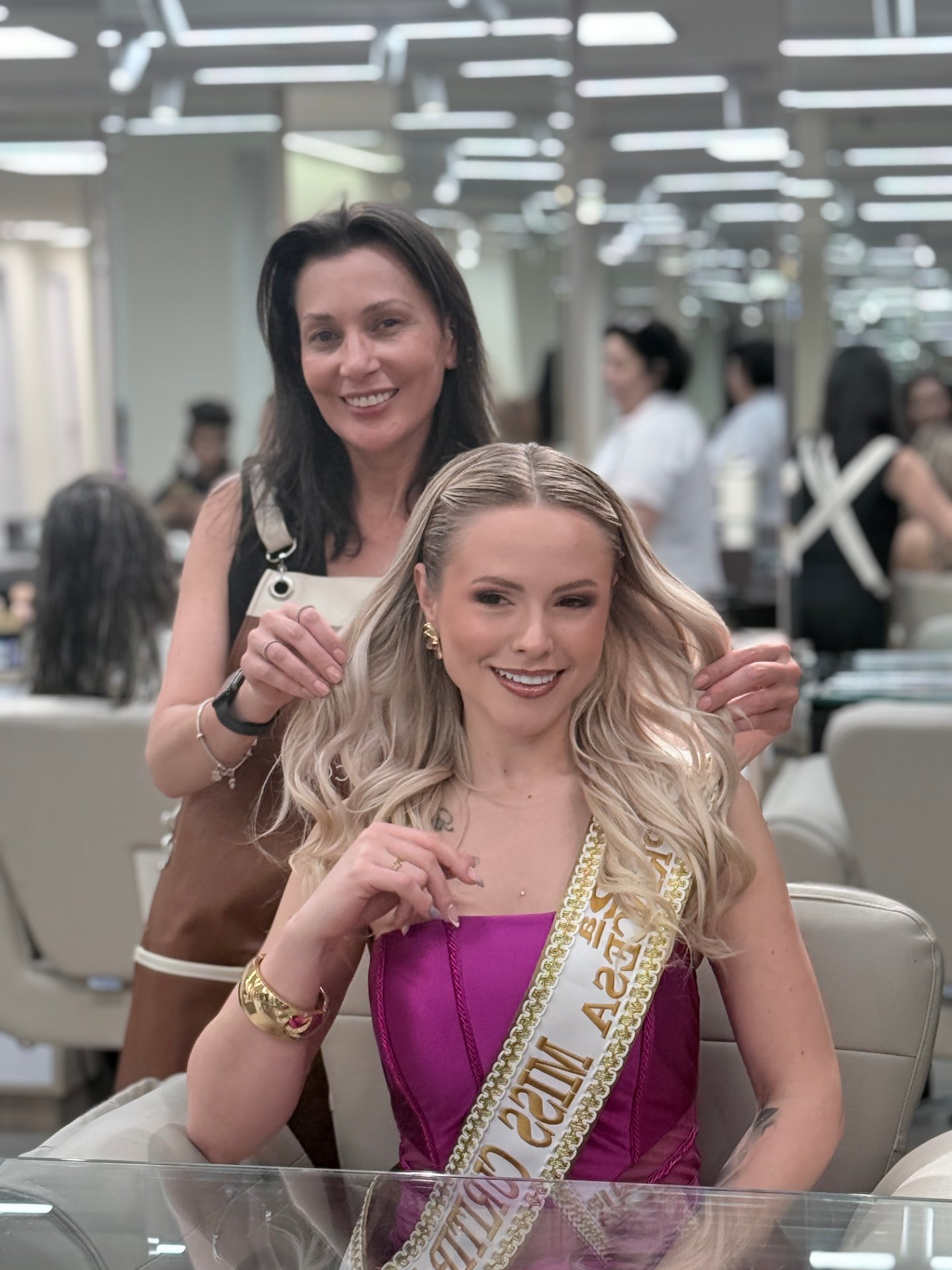 Cliente com faixas de concurso, cabelo longo ondulado em loiro acinzentado e maquiagem, ao lado de profissional do Nix Beauty Salon, no Shopping Palladium, em Curitiba