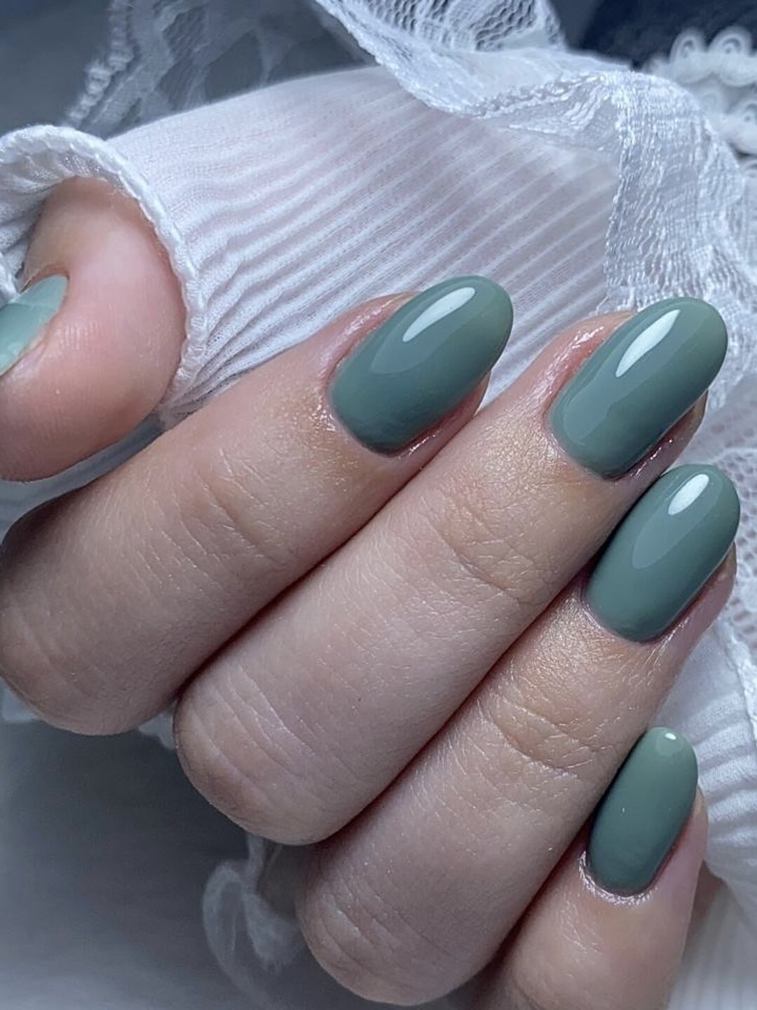 Mãos com unhas em formato oval e esmalte verde sálto brilhante, sobre tecido branco de renda, manicure feita no Nix Beauty Salon, no Shopping Palladium, em Curitiba